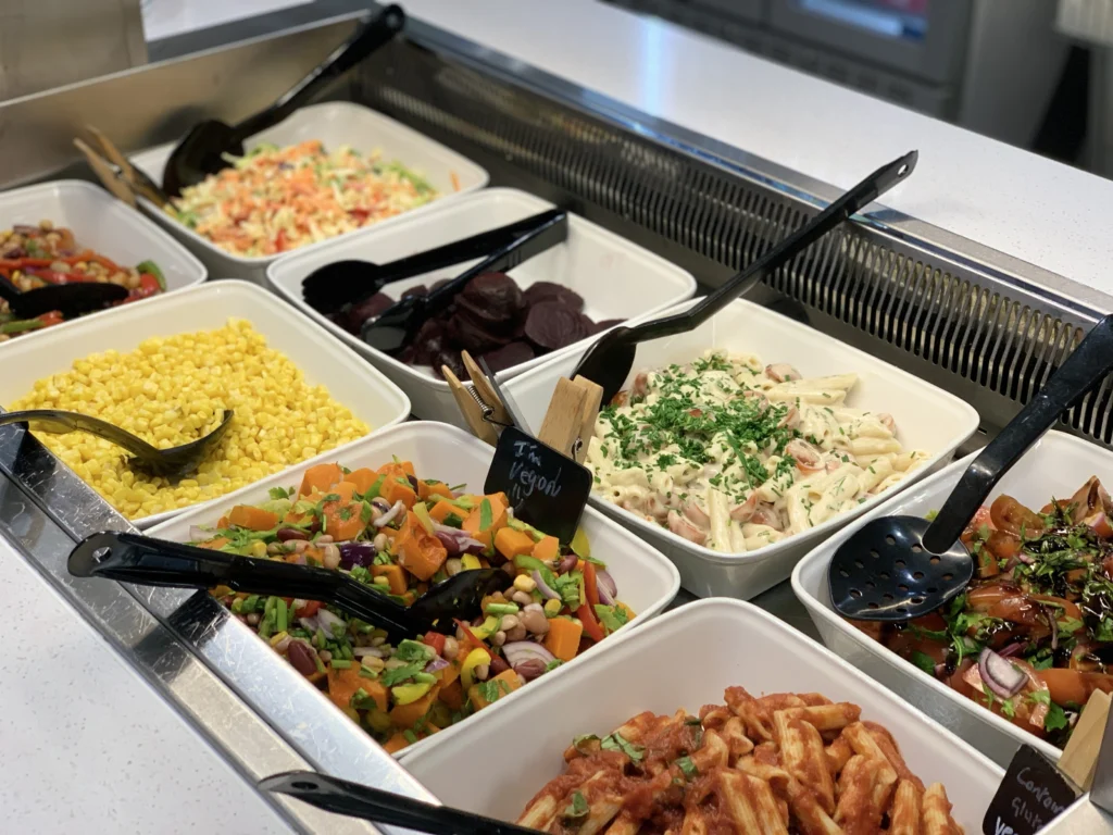 Salad bar at Bistro 32 with a selection of fresh vegetables, toppings, and dressings displayed in stainless steel containers under a glass cover.