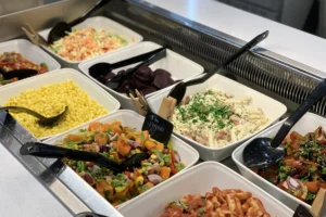 Salad bar at Bistro 32 with a selection of fresh vegetables, toppings, and dressings displayed in stainless steel containers under a glass cover.