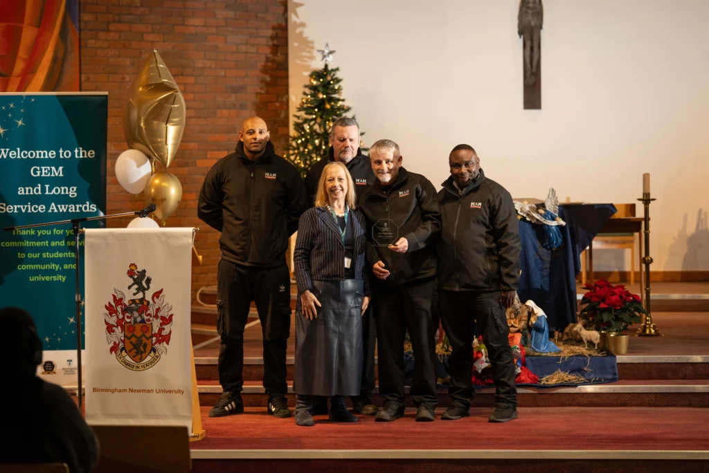 Group of Birmingham Newman University staff standing together at the 2025 Staff Awards ceremony, smiling and holding trophies in front of a branded backdrop.