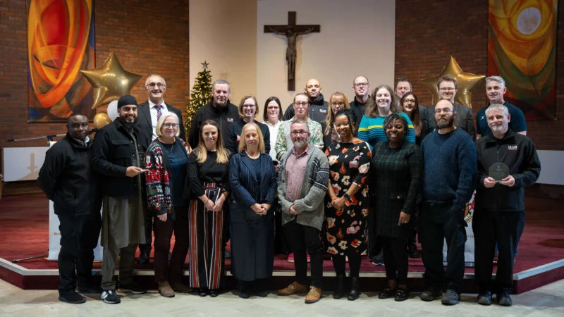 Group of Birmingham Newman University staff standing together at the 2025 Staff Awards ceremony, smiling and holding trophies in front of a branded backdrop.